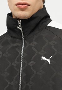 Άνδρας που φοράει μαύρο αθλητικό μπουφάν Puma με ψηλό γιακά, με διακριτικό σχεδιασμό μοτίβου και λευκό λογότυπο Puma στο στήθος.