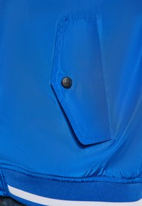 Modrá ľahká bunda s textúrovaným povrchom, bočná kapsa so čiernym gombíkom a pruhovaným rebrovaným lemom na spodnej časti.