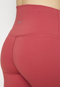Leggings a vita alta, aderenti, in un materiale rosso scuro con cuciture piatte. Presentano un piccolo logo Nike blu vicino alla vita.