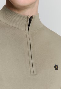 Khaki half-zip trui van ribgebreide stof. Bevat een zwart hexagonaal logo op de borst en een soepele rits met een stoffen trekkoord.