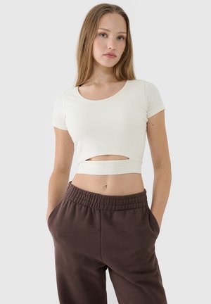 Jonge vrouw die een wit kortemouw cropped top met een uitsnijding aan de voorkant en bruine elastische taillebroeken draagt, staand met handen in de zakken.