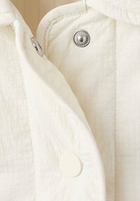 Jakna v smetanasti barvi z mrežnim vzorcem, z okroglim srebrnim sponkam na pritrditev in gladkim smetanastim gumbom. Tkanina je lahka in teksturirana.