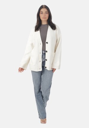 Femme portant un cardigan blanc boutonné, un haut gris ajusté, un jean taille haute bleu clair et des talons ouverts transparents, se tenant contre un fond blanc.