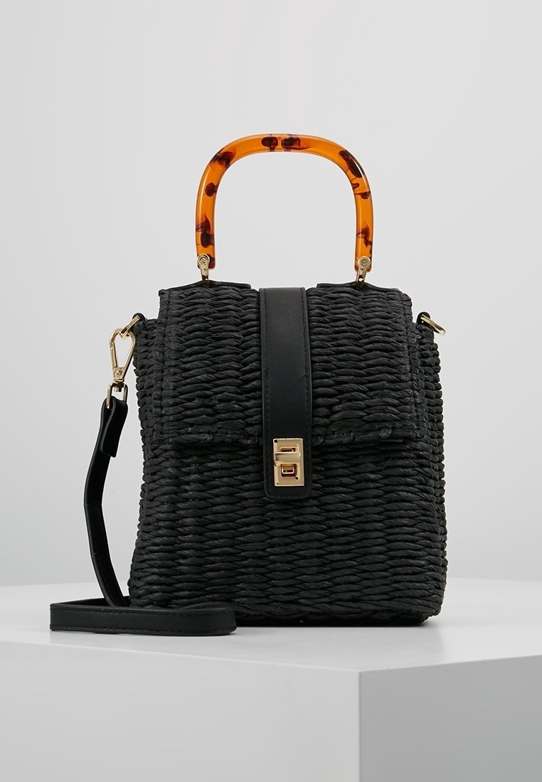 Sac à main tressé noir avec poignée arrondie en écaille de tortue, rabat avant avec fermoir doré et bandoulière amovible sur surface blanche.