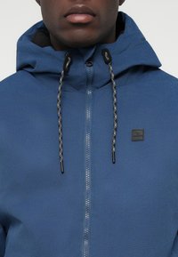 Volwassene in een blauw gewatteerd jack met rits, voorzien van zwarte en grijze trekkoorden en een klein zwart logo op de borst.