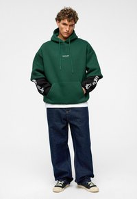 Haut à capuche vert avec le logo "IMPACT", manches noires avec des graphiques blancs, associé à un jean bleu ample et des baskets noires. Matériau en coton, coupe décontractée.