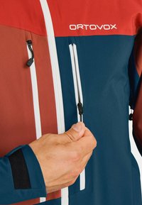 Hand adjusting chest zipper on blue and red Ortovox jacket with white zipper details and black wrist patch.