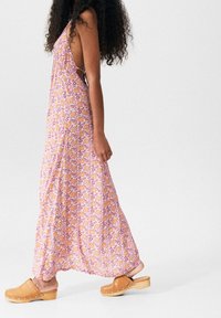 Vestido maxi en rosa con estampado de flores amarillas, que presenta una espalda profunda y una silueta fluida; combinado con zuecos beige con suelas de madera.