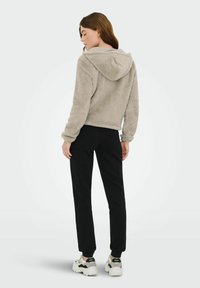 Sweat à capuche en polaire gris clair avec une texture douce, coupe courte et capuche, porté avec un pantalon noir et des baskets blanches avec des accents noirs.