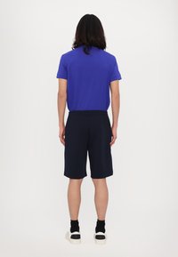 Camicia azzurra a maniche corte con una vestibilità dritta, abbinata a pantaloni corti blu navy. Il modello indossa sneakers bianche con dettagli neri. Design minimalista.
