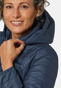Veste matelassée bleu marine avec une surface texturée. Dotée d'une capuche, d'une fermeture éclair et d'un design matelassé, montrant un ajustement confortable. Main près du col.