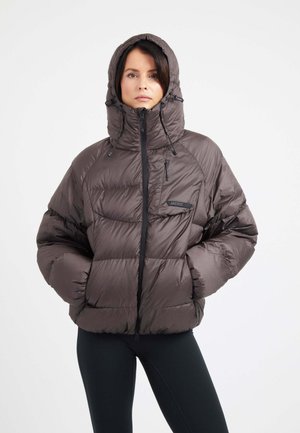 Mujer con una chaqueta acolchada marrón con capucha puesta, manos en los bolsillos, de pie delante de un fondo claro y liso.