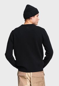 Čierny pletený sveter s rebrovanou textúrou, zakriveným výstrihom a štítkom s logom na zadnej strane. Kombinovaný s svetlými nohavicami.