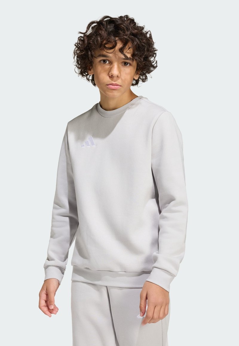 Adolescente con cabello rizado que lleva una sudadera Adidas gris claro y pantalones a juego, de pie y mirando a la cámara sobre un fondo liso.