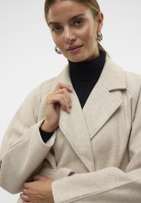 Cappotto di lana beige con colletto a scialle, design avvolgente e maniche ampie. Indossato su un dolcevita nero, con una mano che tiene il colletto.