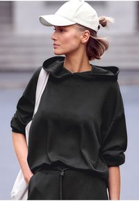 Čierna mikina s kapucňou, voľný strih a obrolené rukávy, v kombinácii s nohavicami na šnúrku. Svetlo béžová čiapka dotvára celý outfit.