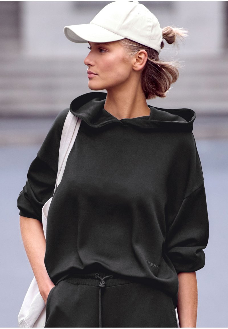 Čierna mikina s kapucňou, voľný strih a obrolené rukávy, v kombinácii s nohavicami na šnúrku. Svetlo béžová čiapka dotvára celý outfit.