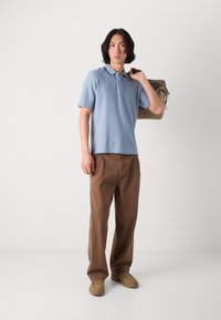 Hellblaues Poloshirt mit kurzen Ärmeln, beige Tasche über der Schulter, braune locker sitzende Hose und beige Schlupfschuhe vor einem schlichten Hintergrund.
