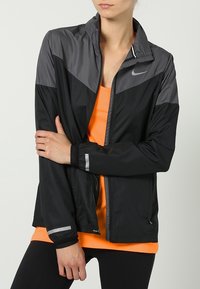 Nike letvægts vindjakke med sorte og grå farveblokke, lynlås foran, reflekterende detaljer og elastiske manchetter over en orange top.