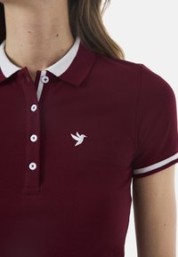Burgundi polo särk valgete triipudega kraega, millel on viie nupuga suletus ja väike brodeeritud linnulogo vasakul rinnaosas.