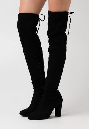 Czarne zamszowe kozaki za kolano na wysokim obcasie, z zaokrąglonymi noskami i wiązaniami z tyłu, noszone na gołych nogach.