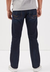 Jeans de mezclilla azul oscuro con un corte relajado, con dos bolsillos traseros que presentan costuras amarillas, llevados con una camiseta gris y zapatillas blancas.