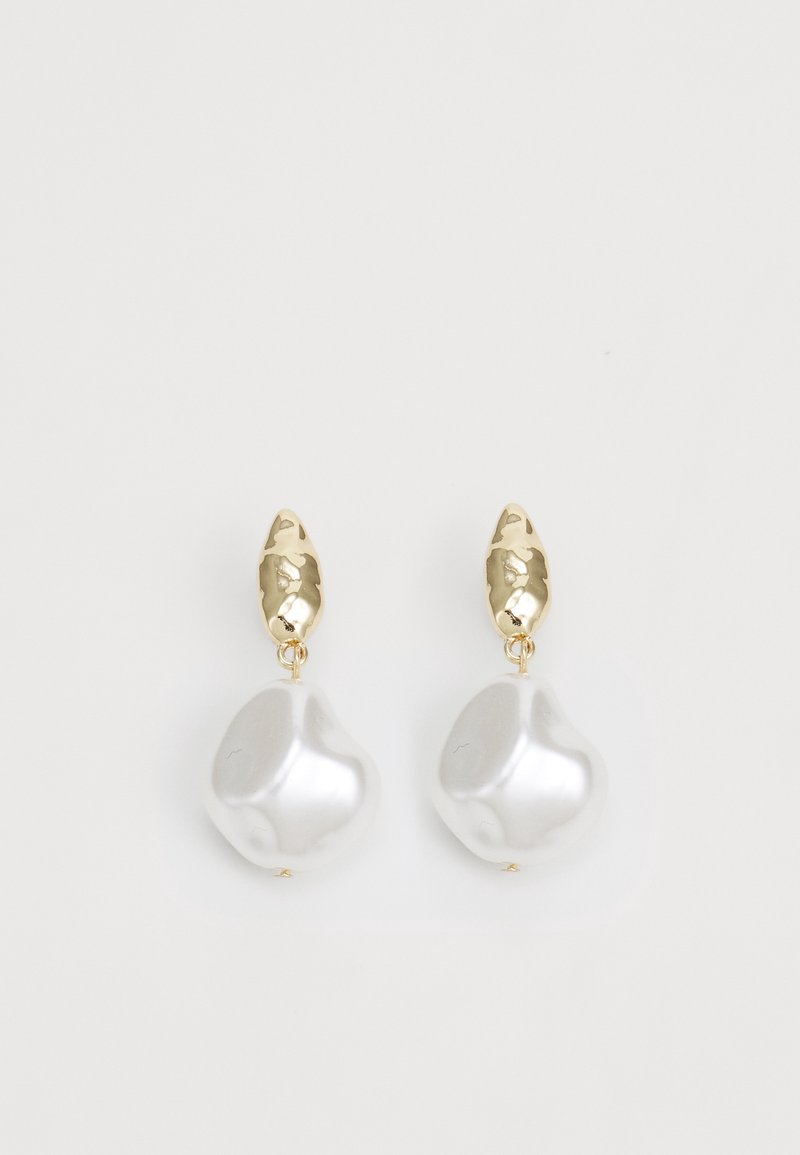 Boucles d'oreilles clous martelées dorées avec grands pendentifs goutte perlée blanche de forme irrégulière sur fond blanc.