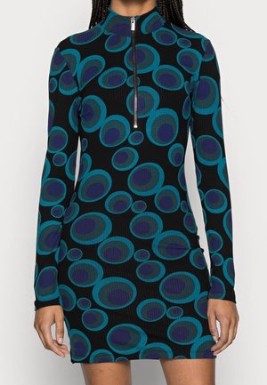 Langärmeliges schwarzes Kleid mit türkisblauen und marineblauen kreisförmigen Mustern, einem hohen Kragen und einem Frontreißverschluss, getragen von einer Person mit geflochtenem Haar.