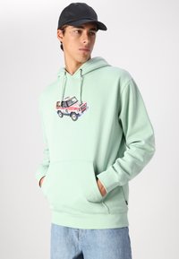 Mentolová mikina s kapsou na přední straně, která má barevnou grafiku vintage auta. Měkká tkanina a nastavitelná kapuce se šňůrkou. Volný střih.