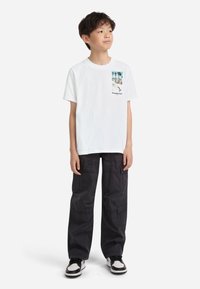 Balta kokvilnas T-krekls ar drukātu fotoattēlu un tekstu priekšpusē, apvienots ar brīvi pielāgotām melnām kravas biksēm ar kabatām un strukturētu virsmu.