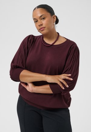 Femme aux cheveux foncés en chignon porte un pull bordeaux à manches bouffantes, un collier de perles assorti et un pantalon noir, bras croisés.