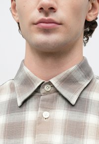 Detailný záber dolnej časti tváre a krku osoby, ktorá má na sebe bežovo-šedú karovanú košeľu na gombíky s špicatým golierom.