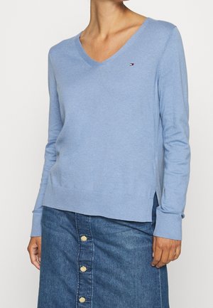 Žena v světle modrém svetru s výstřihem do V s malým logem na hrudi a modré džínové sukni s knoflíky vpředu, stojící před bílým pozadím.