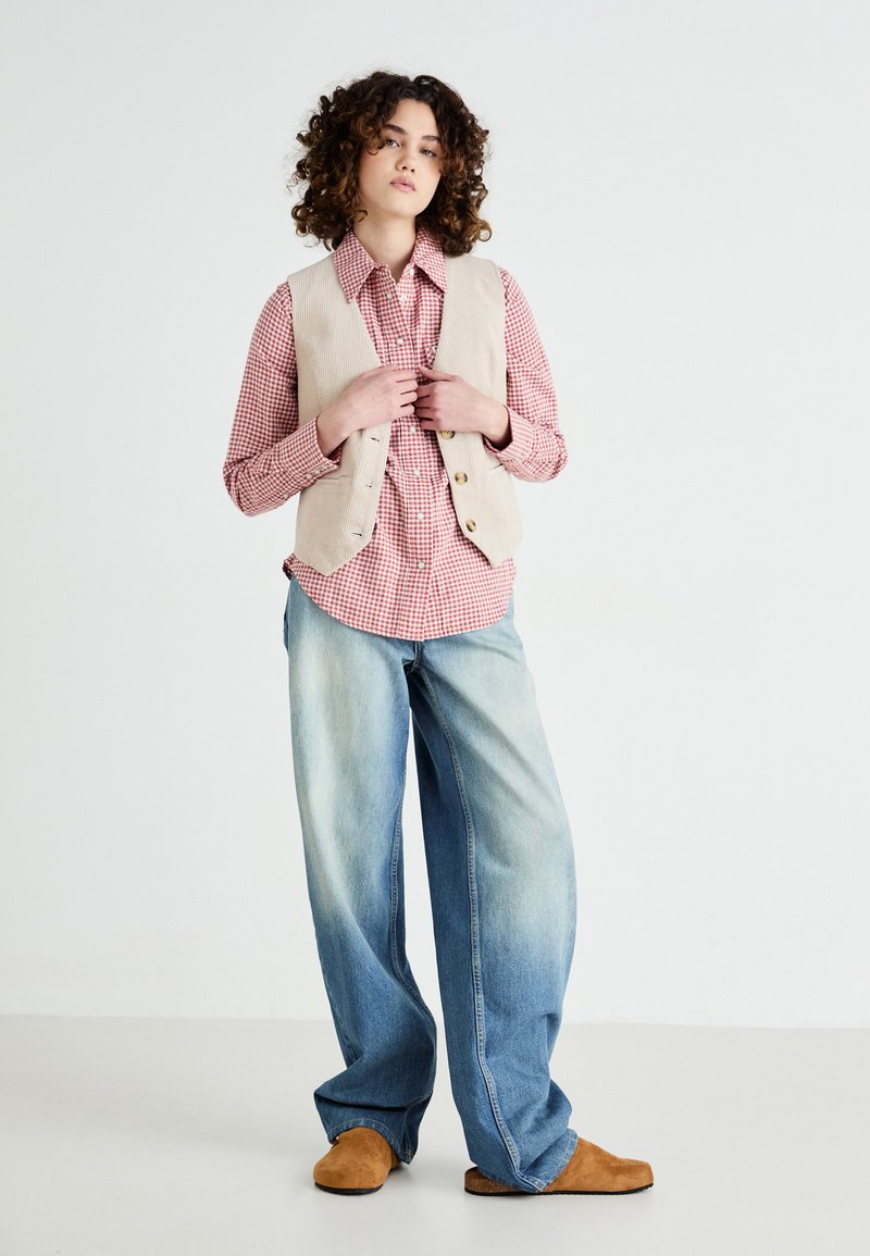 Beige Corduroy-Weste über einem rot-weißen Karohemd, kombiniert mit übergroßen hellblauen Jeans und braunen Wildlederschuhen. Schlichter weißer Hintergrund.