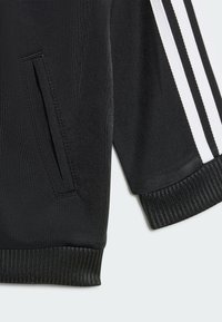 Black athletic jacket featuring textured fabric, zippered side pocket, and white striped accents on the sleeves. Ribbed cuffs and hem.