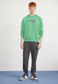 Sweatshirt verde com um logotipo redondo, combinado com calças de treino cinzentas e ténis preto e branco. Tecido liso com punhos canelados.