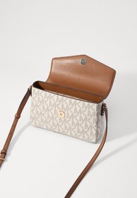 Beige crossbody taske med et gentagende logomønster, brune læderdetaljer, en trykknaplukning og et justerbart skulderrem. Tekstur- og struktureret design.