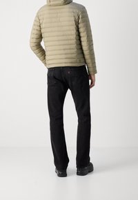 Helle olivfarbene Pufferjacke mit horizontaler Steppung, abnehmbarer Kapuze und elastischen Bündchen, kombiniert mit schwarzen Jeans und schwarzen Schuhen.