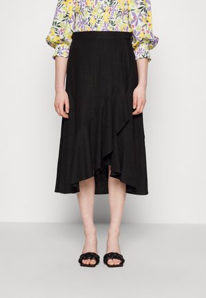 Vrouw draagt een zwarte asymmetrische midi-rok met ruches, een bloemenblouse met lange mouwen en zwarte gevlochten sandalen met open tenen.
