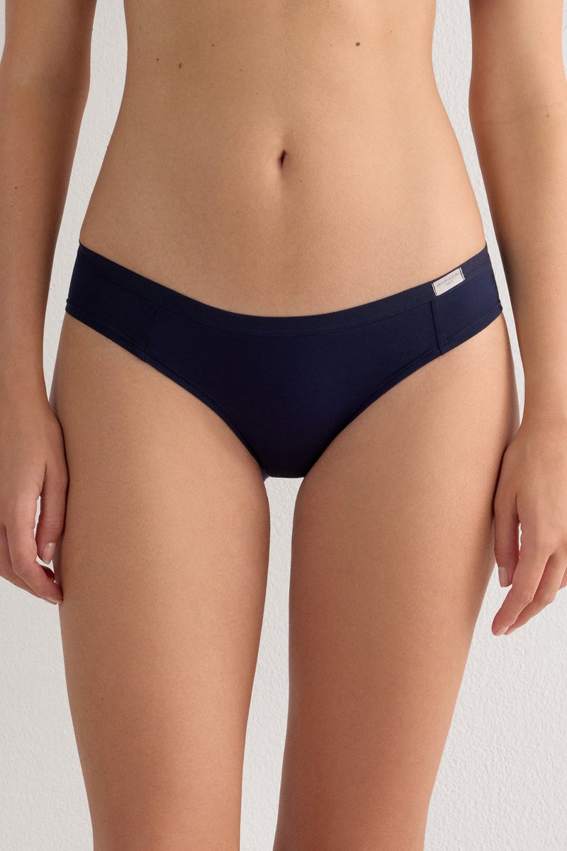 Frau trägt einfache dunkelblaue Bikini-Unterwäsche, ab Beschnitt von Brust bis oberem Oberschenkel vor hellem Hintergrund.