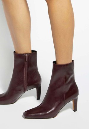 Bottines en cuir marron à bouts pointus, fermetures éclair latérales et talons de hauteur moyenne portées sur des jambes nues sur fond blanc.