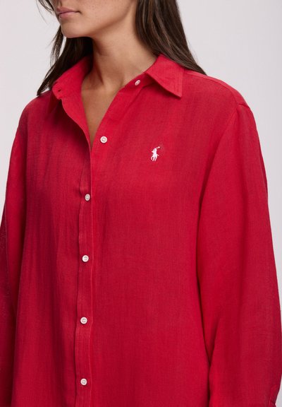 Κόκκινο πουκάμισο από λινό με γιακά, μακριά μανίκια και λευκά κουμπιά στο μπροστινό μέρος. Διαθέτει μικρό λευκό λογότυπο στο αριστερό στήθος.