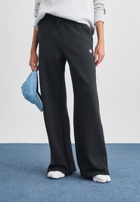 Černé tepláky s volnějším střihem, elastickým pasem a malým logem. Doplněné světle modrou čepicí drženou v levé ruce.