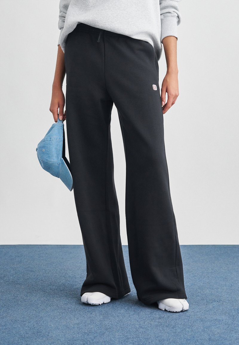 Černé tepláky s volnějším střihem, elastickým pasem a malým logem. Doplněné světle modrou čepicí drženou v levé ruce.