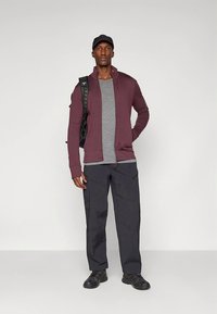 Tumši sarkans apģērbs ar rāvējslēdzēju virs pelēka krekla, kombinēts ar melnām darba biksēm. Uz kreisā pleca mugursoma, melni sporta apavi un cepure. Vienkāršs dizains.