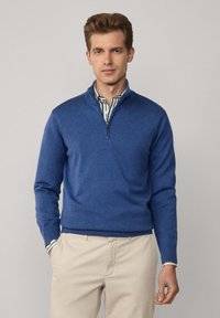 Pull bleu à col zippé en tricot doux, avec des manches longues et des poignets côtelés. Porté par-dessus une chemise à col rayée.