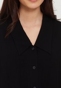 Schwarze Hemd mit Knopfleiste, das einen großen Kragen, eine glatte Textur und vier Knöpfe an der Vorderseite aufweist und sich auf ein sauberes, minimalistisches Design konzentriert.