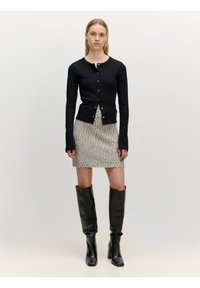 Cardigan noir à manches longues avec des boutons, associé à une mini-jupe grise texturée. Le modèle porte des bottes en cuir noir jusqu'aux genoux.
