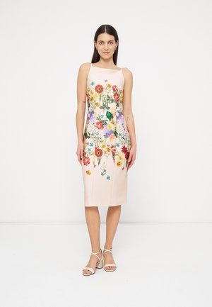 Femeie purtând o rochie midi fără mâneci, de culoare roz pal, cu imprimeu floral colorat și sandale albe cu toc, stând în picioare pe un fundal alb simplu.