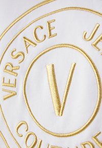 Vitt tyg med en framträdande guldbroderad logotyp med "Versace Jeans Couture" och ett centralt stiliserat "V". Cirkulär design detalj.